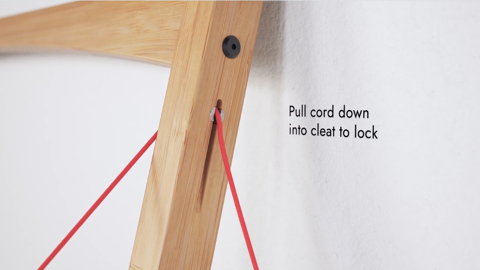 Close-up of wooden desk frame showing red cord and cleat mechanism, with instructional text: "Pull cord down into cleat to lock."