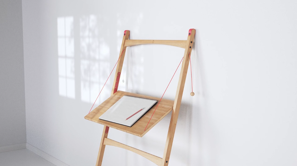 Minimalist wooden wall-mounted desk with red support cords, holding a sketchpad and pencil, positioned in a sunlit room with shadows from a window on a white wall.