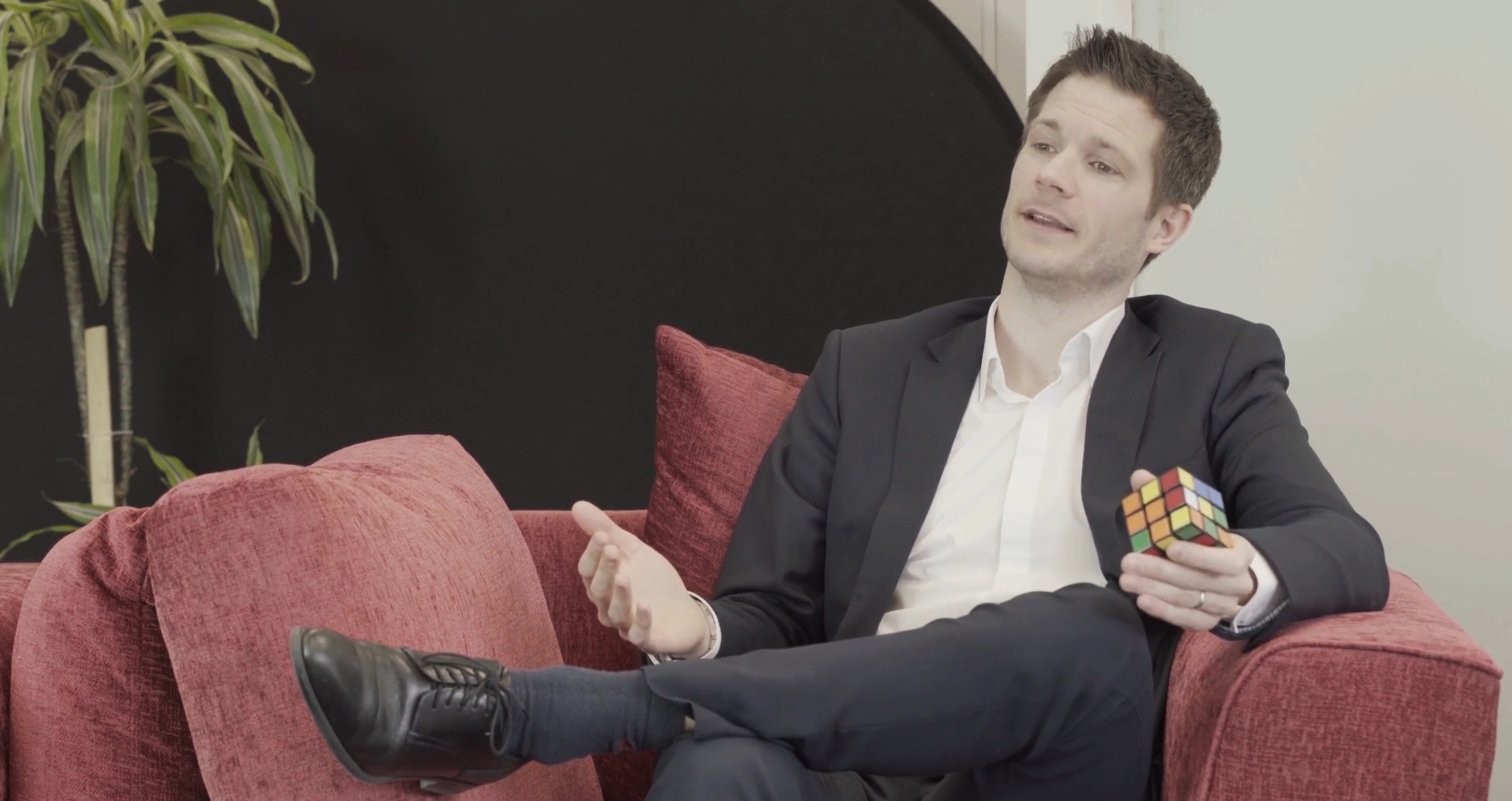Man in a suit sitting on a red sofa holding a Rubik’s Cube, speaking with animated gestures, with a plant and black backdrop in a relaxed lounge-style setting.