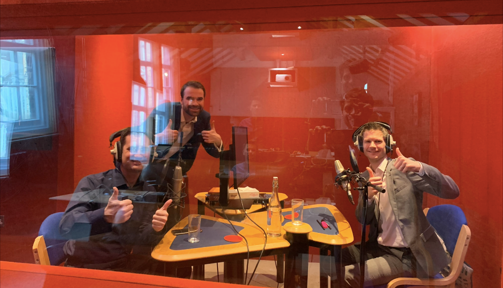 Three men recording in a professional sound booth, wearing headphones and giving thumbs up, surrounded by microphones, a table with glasses, and a red-walled studio background.
