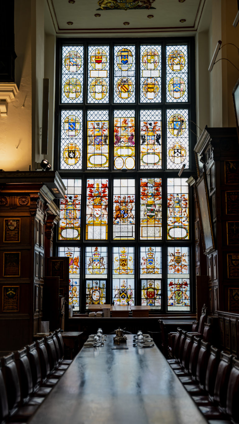 Historic dining hall with long wooden table and high-backed chairs, featuring an ornate stained-glass window displaying colorful heraldic crests, lit by natural light.