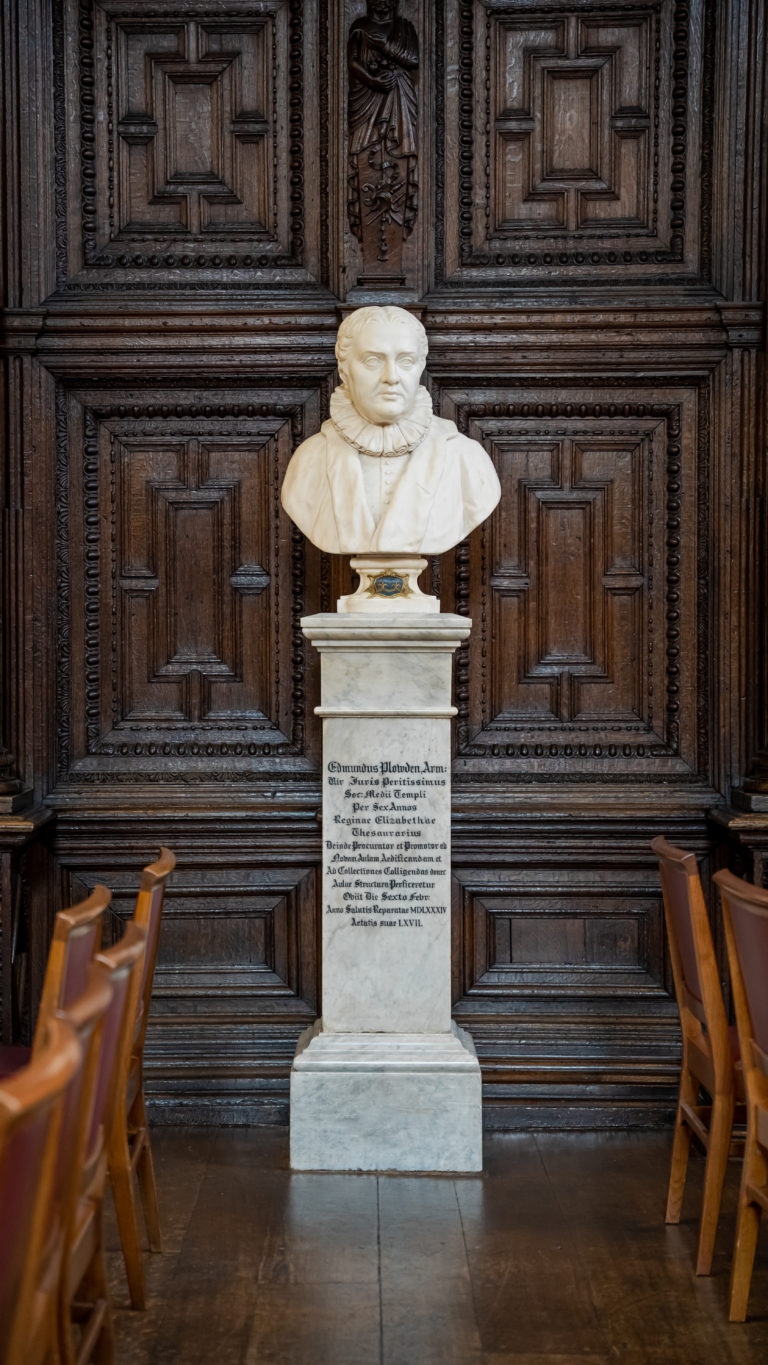 Marble bust of Edmund Plowden displayed on a pedestal with Latin inscription, set against a richly carved wooden wall in a formal hall with arranged wooden chairs.