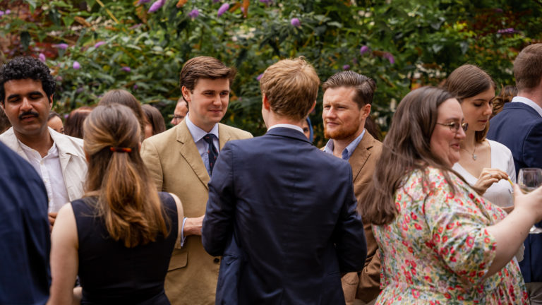 Students dressed in formal and semi-formal attire chatting and enjoying drinks at an outdoor event surrounded by lush greenery and blooming flowers.