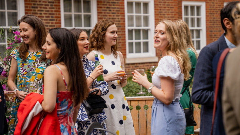 Group of students socialising and holding drinks at an outdoor event in a courtyard with brick walls and white-framed windows in the background.