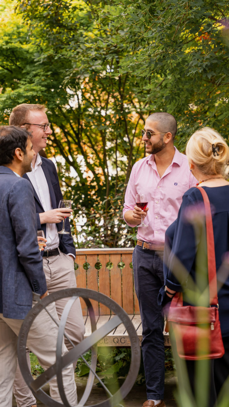Law students socialising outdoors in a leafy garden, holding glasses of wine and engaged in conversation near a wooden bench and decorative sundial.