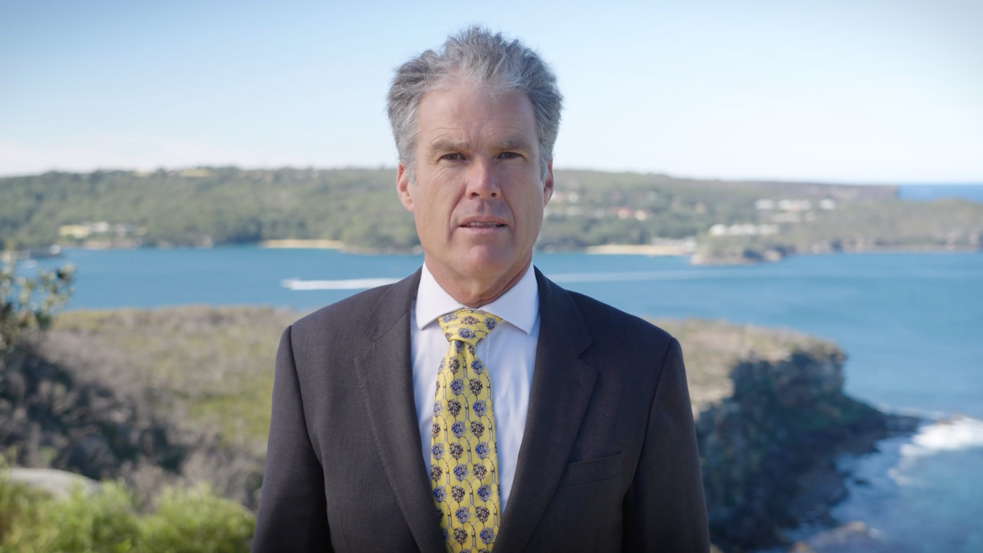 Man in a suit with a yellow patterned tie speaking outdoors, with a scenic coastal background of blue waters and green hills.