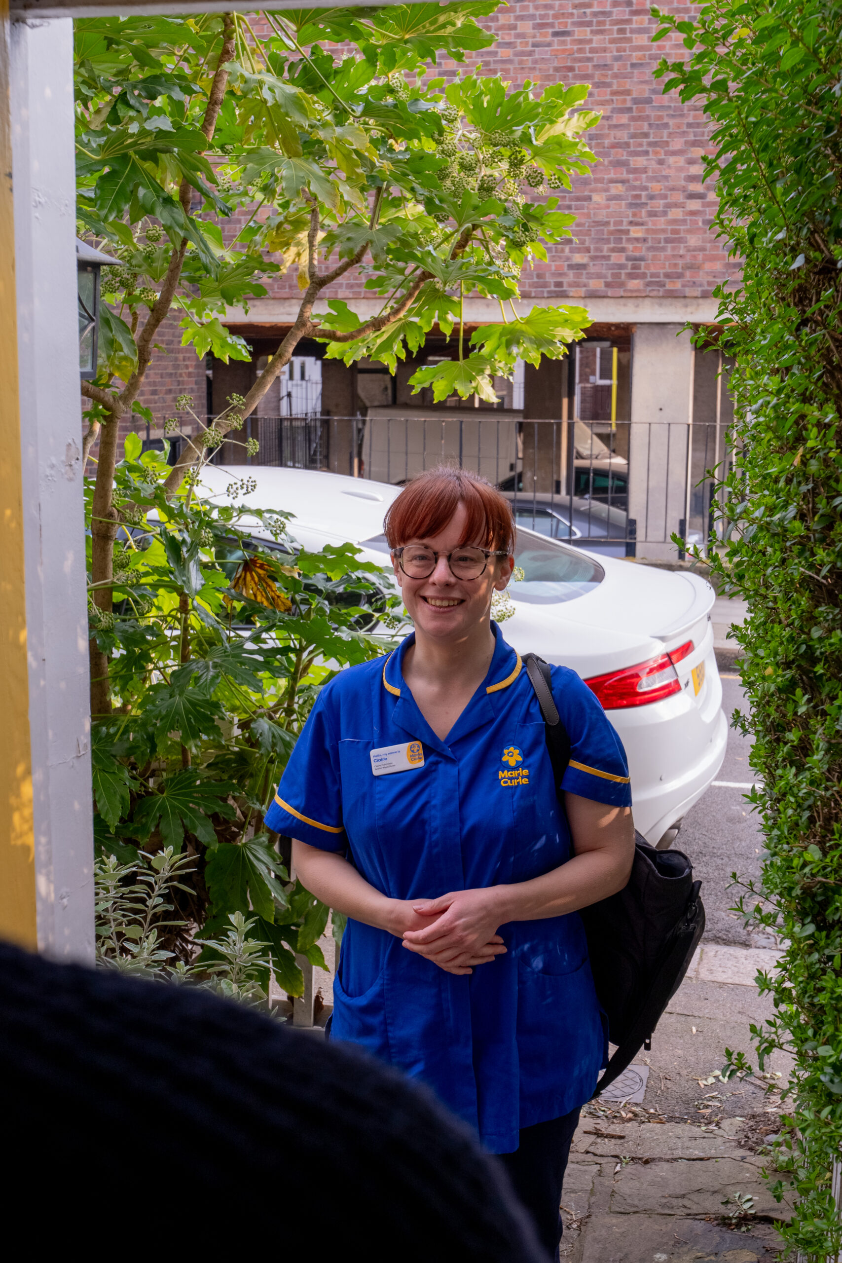 Marie Curie nurse in uniform smiling at the doorway during a home visit, standing outside near a white car and leafy green plants.