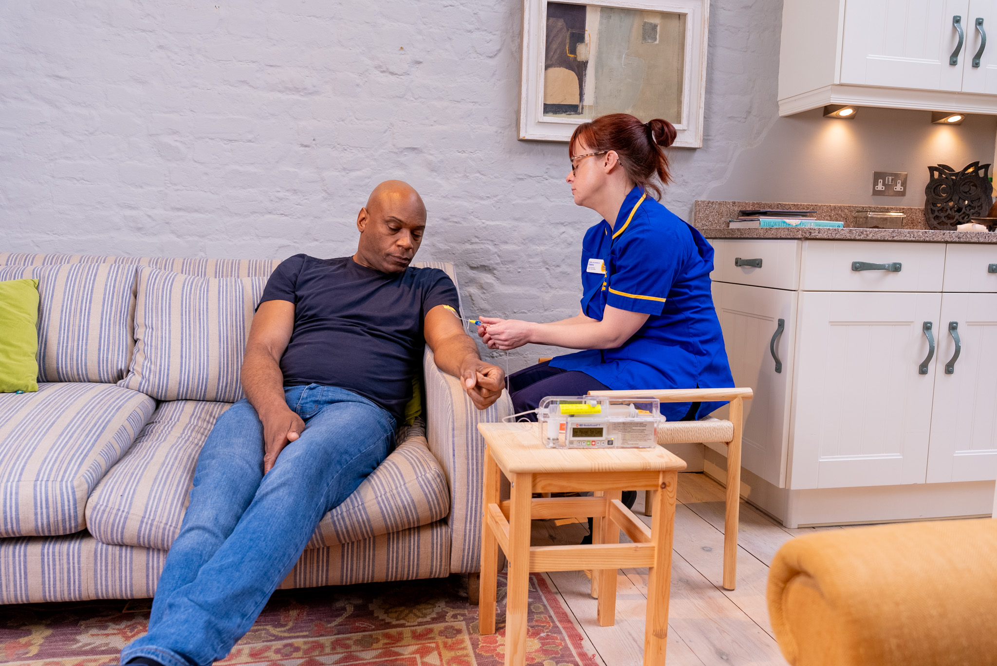 Marie Curie nurse administering syringe driver medication to a patient seated on a sofa in a home setting, with medical equipment placed on a wooden table nearby.