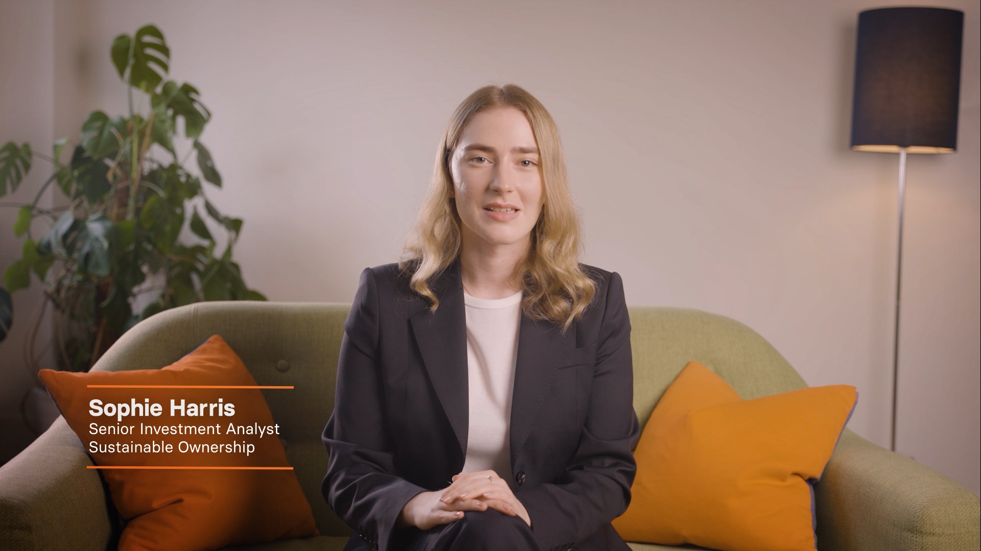 Sophie Harris, Senior Investment Analyst for Sustainable Ownership, sits on a green sofa with orange cushions, delivering an interview in a warmly lit room with a houseplant and lamp.