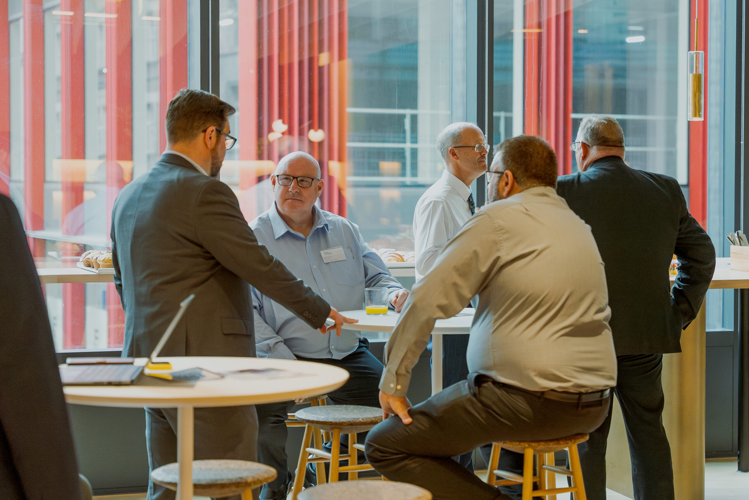 Group of men in business attire engaged in conversation around tall tables by large windows during a professional conference break.