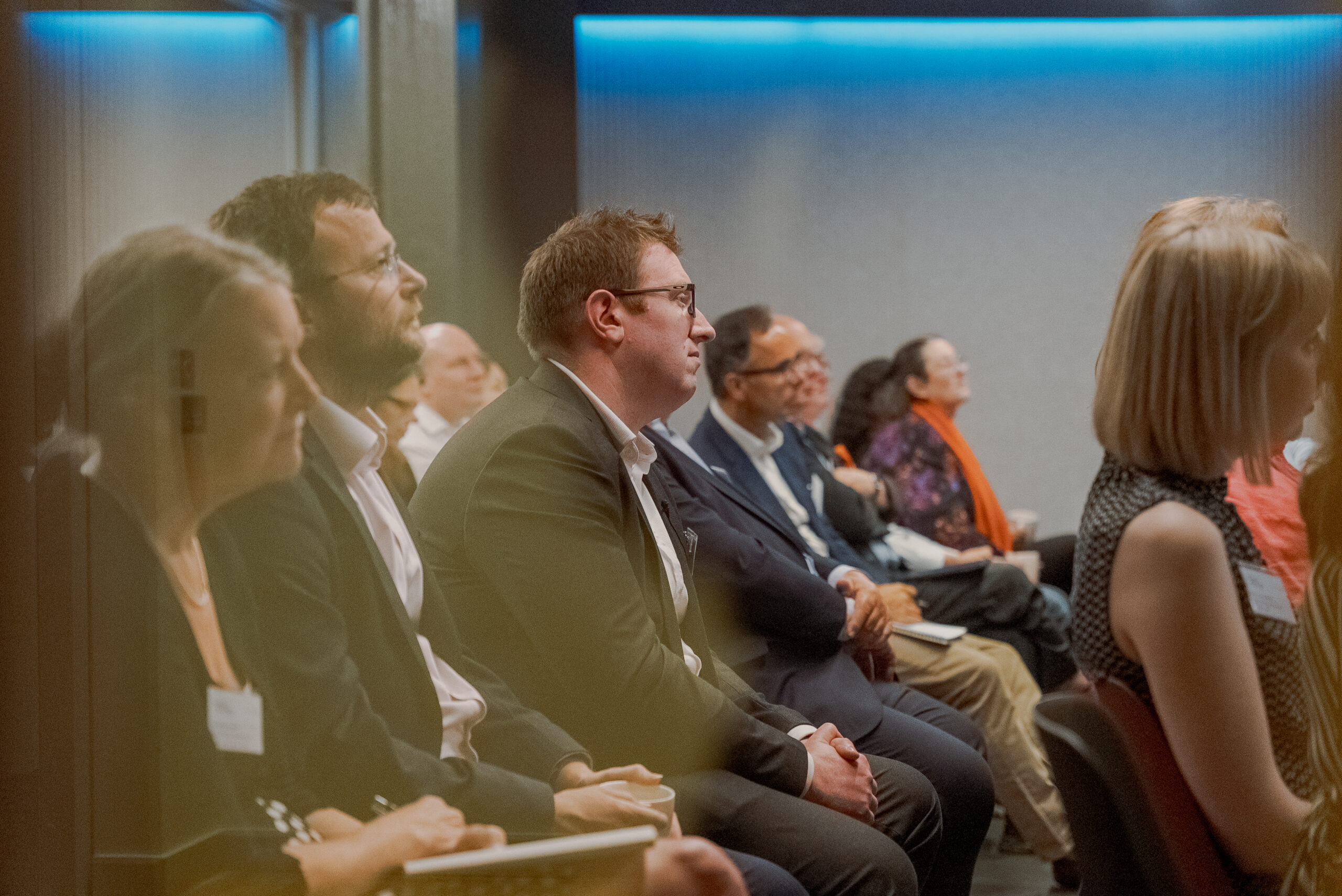 Audience members seated and attentively listening during a conference session in a dimly lit room.