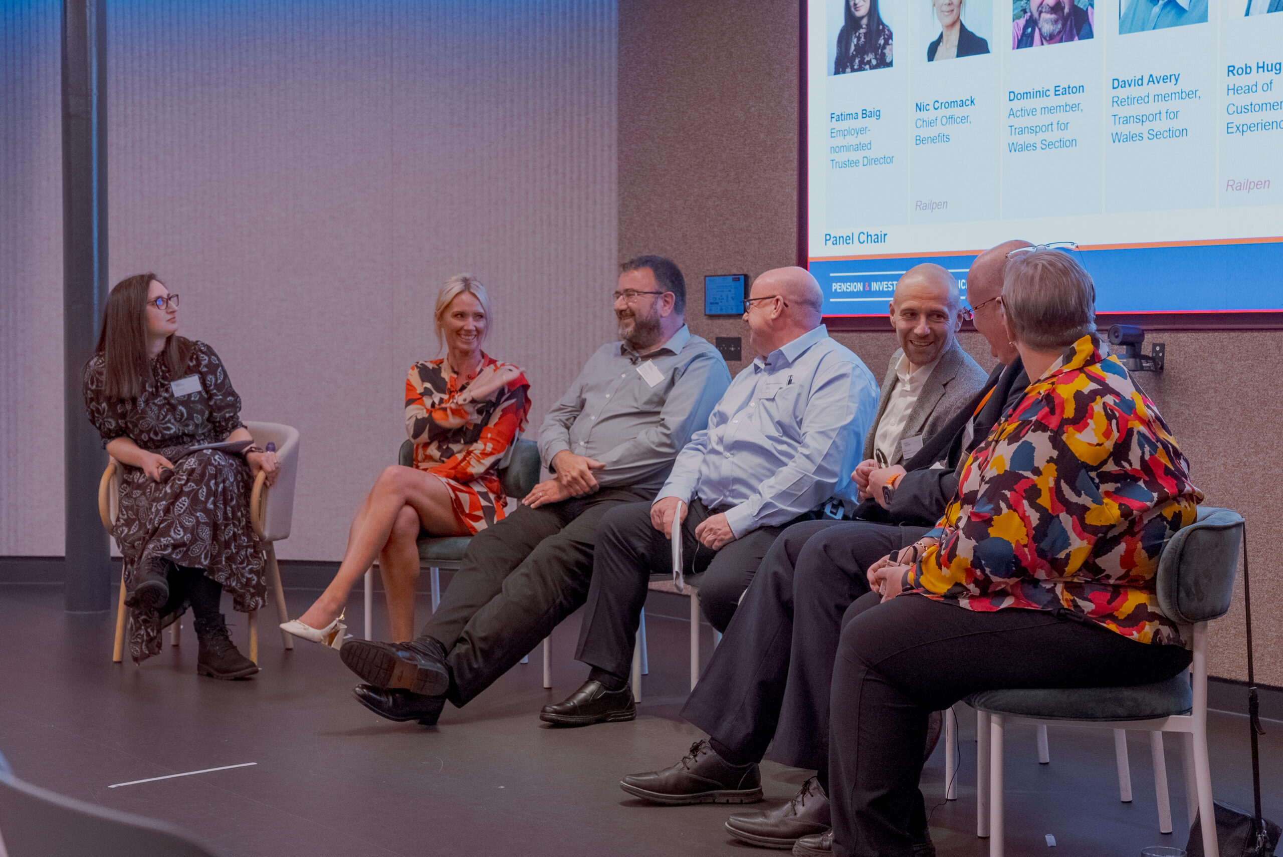 Panel of speakers seated on stage during a conference, engaging in discussion with a projected slide listing their names and roles.