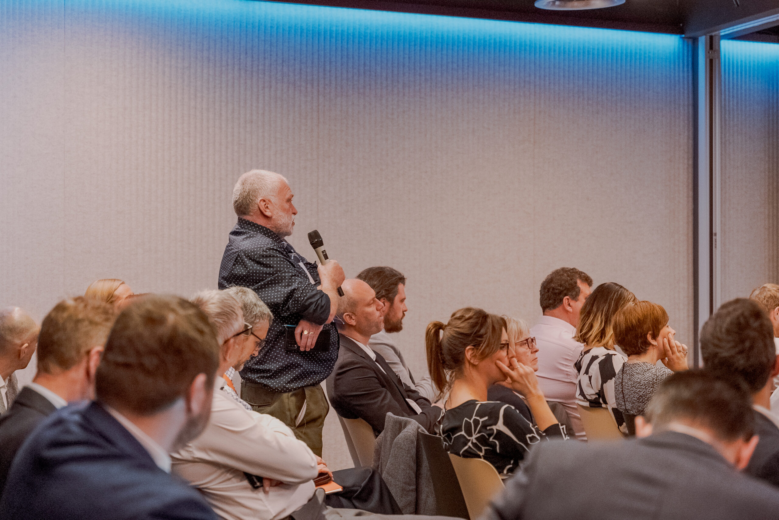 Man standing and speaking into a microphone during a conference session, surrounded by seated attendees listening attentively.