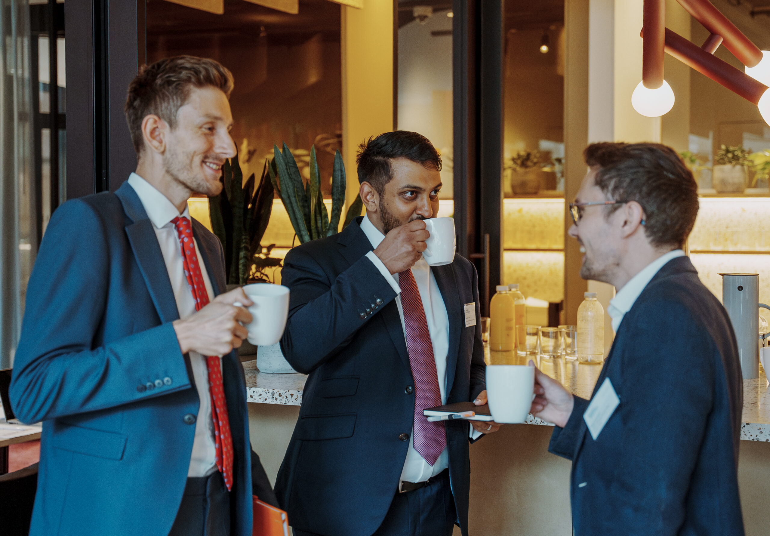 Three professionally dressed men enjoy coffee and conversation in a modern conference lounge with warm lighting and a refreshments counter.