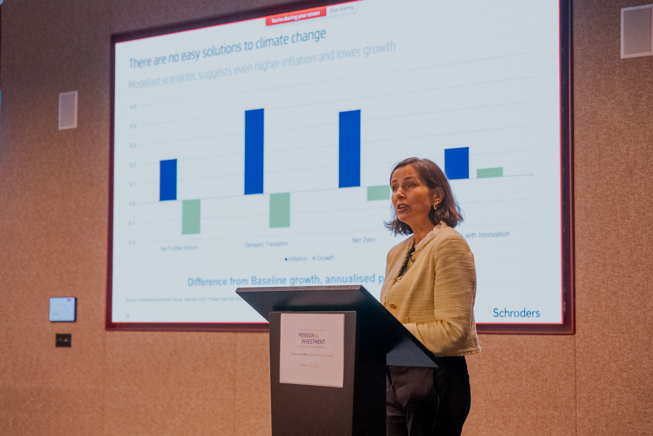 A woman presents at a climate-focused conference, standing at a podium in front of a projected bar graph comparing inflation and growth across climate scenarios.