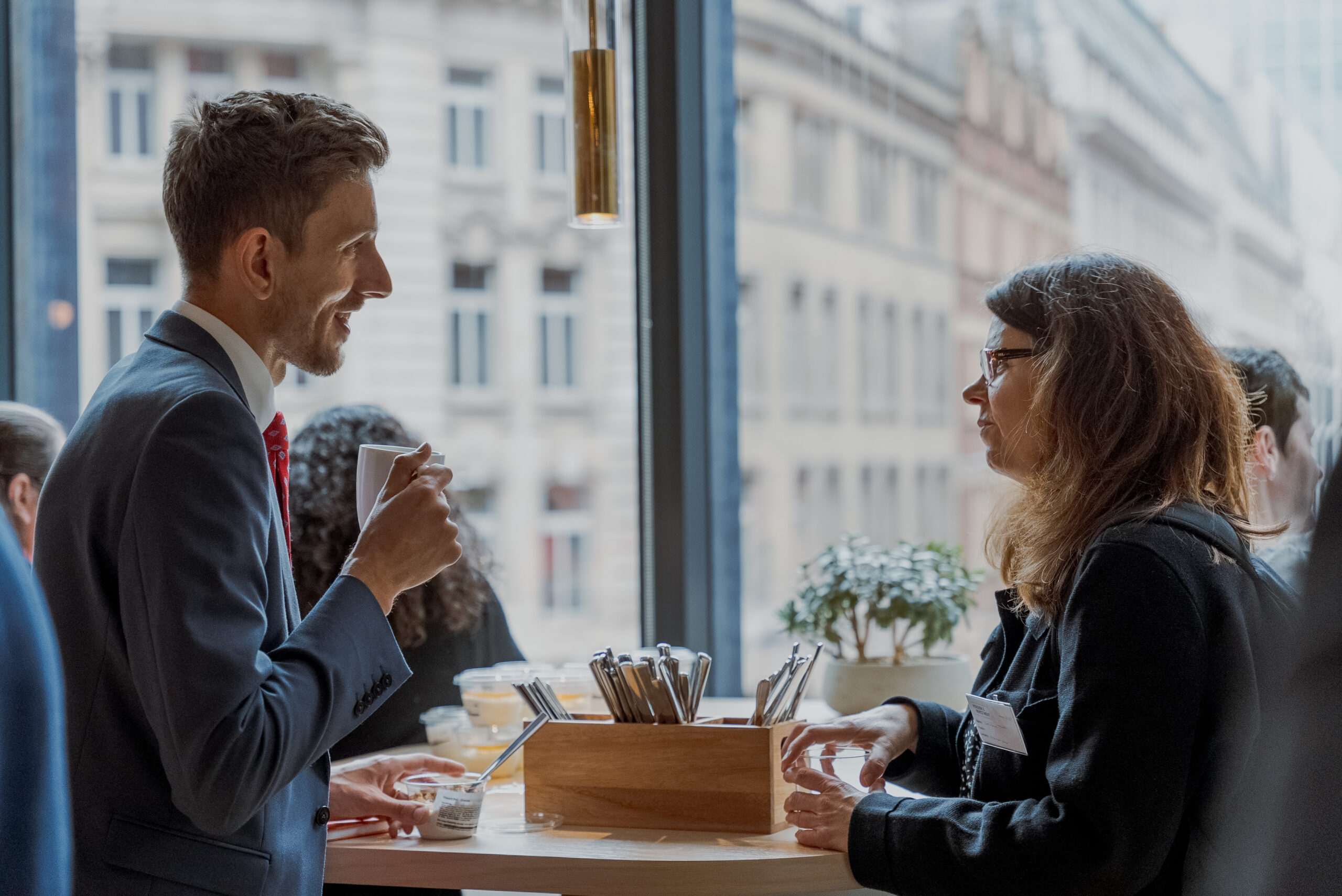 A man and woman in business attire chat over coffee and snacks at a tall table by a large window overlooking a city street.