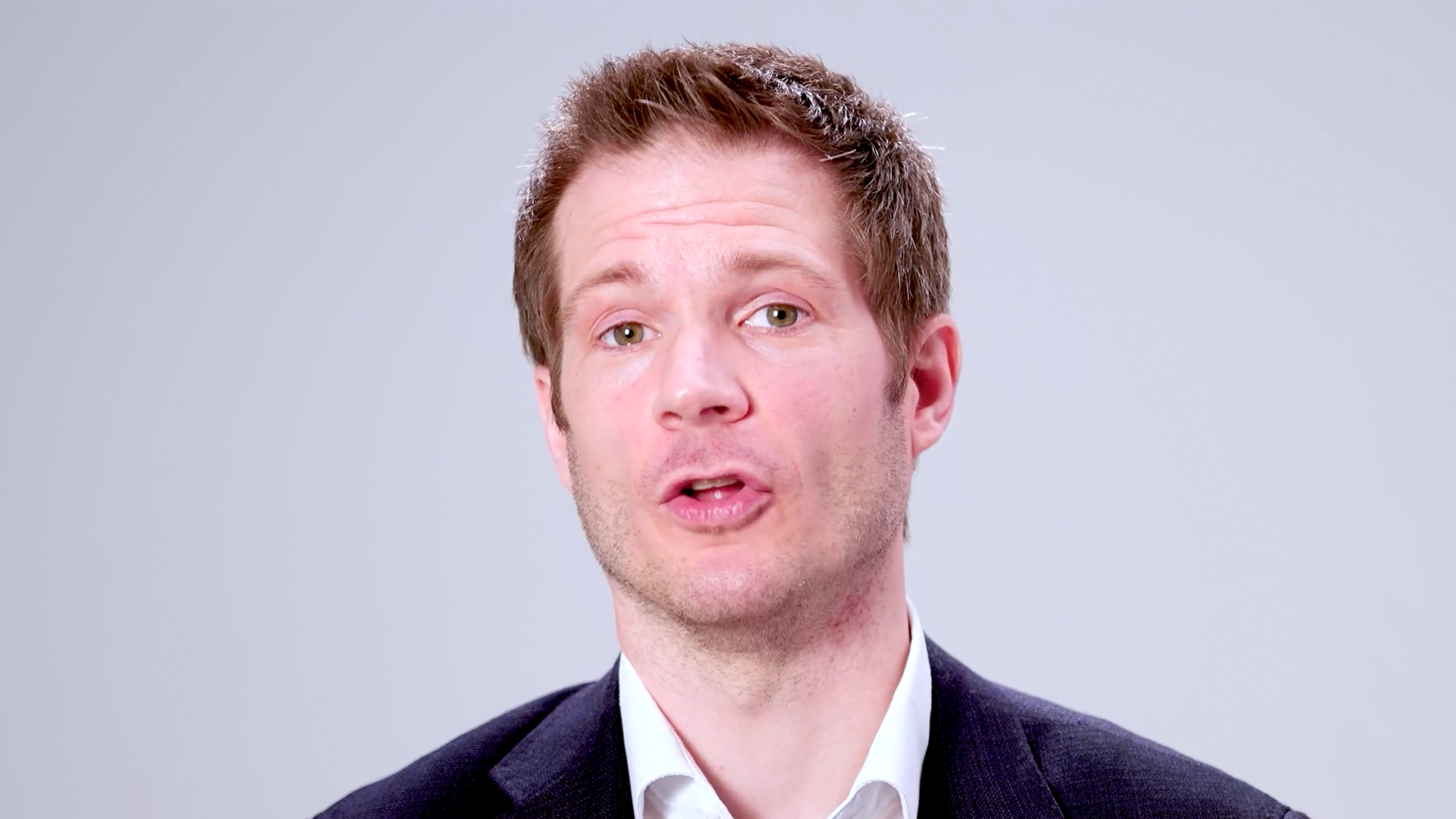 Close-up of a man in a suit speaking directly to the camera against a plain studio background, likely presenting or explaining a financial or business topic.