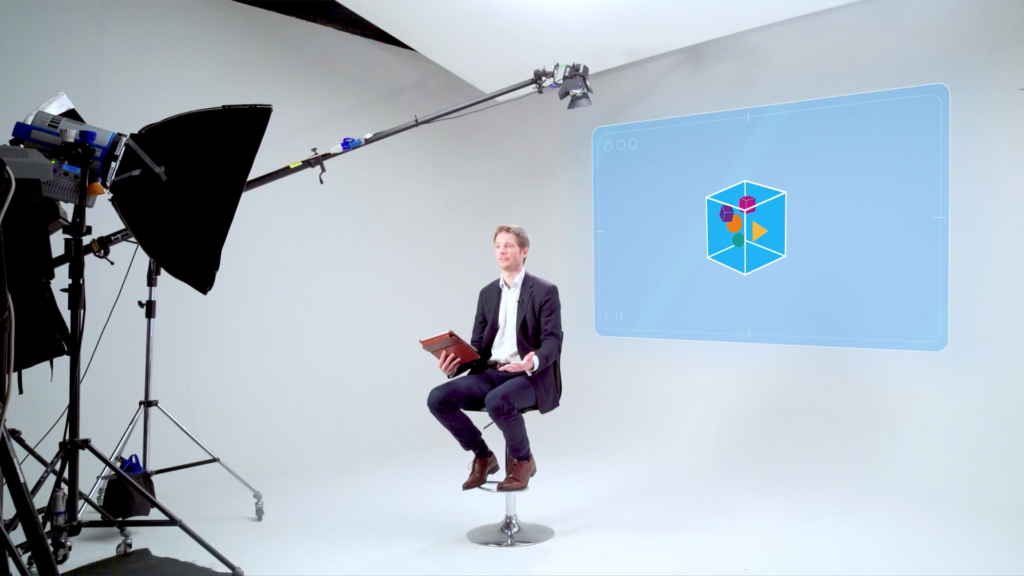 Man in a studio setting sits on a stool with tablet in hand, speaking beside a large screen displaying a colorful 3D cube graphic, surrounded by professional lighting equipment.