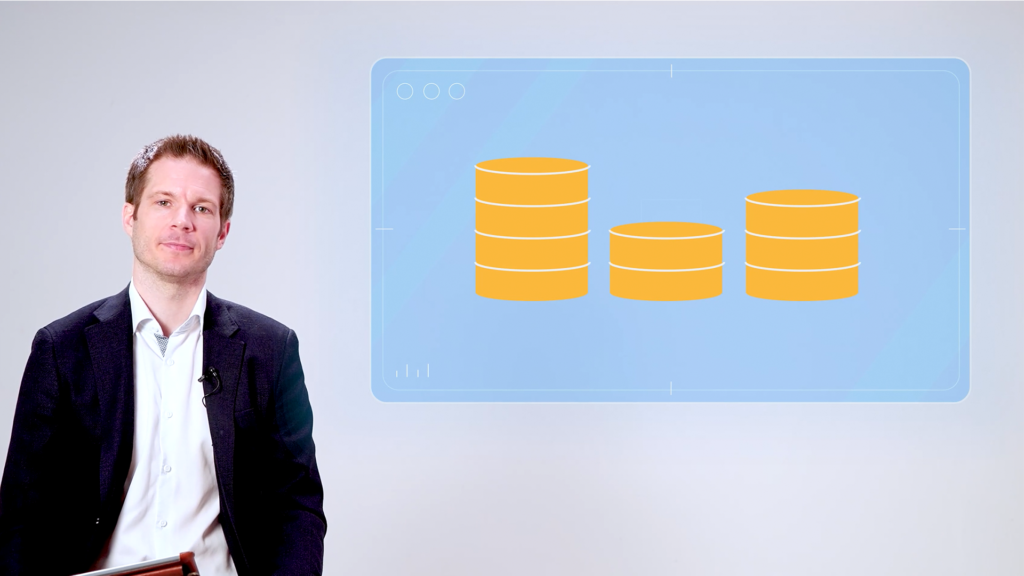 Man in business attire sits beside a digital screen displaying three stacks of gold coins, illustrating financial or investment concepts in a studio presentation setting.