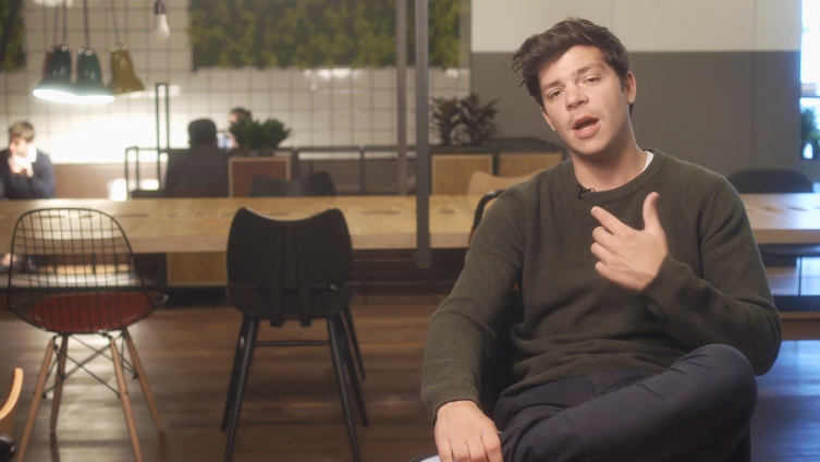 Man speaking while seated in a modern WeWork office space, with a large wooden table and stylish chairs in the background.