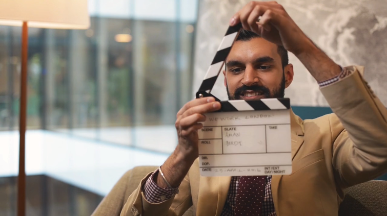 Man in a beige blazer holding a clapperboard, preparing for a video shoot in a modern office setting with large windows and stylish interior decor.