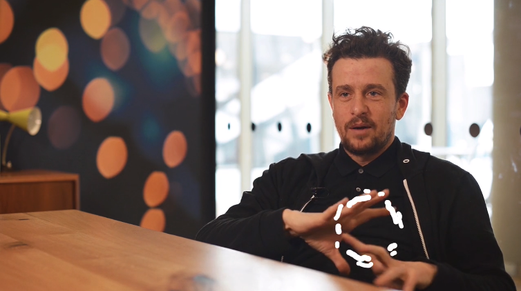 Man speaking at a wooden desk in a modern office with bokeh light artwork and large windows in the background, mid-gesture with a thoughtful expression.