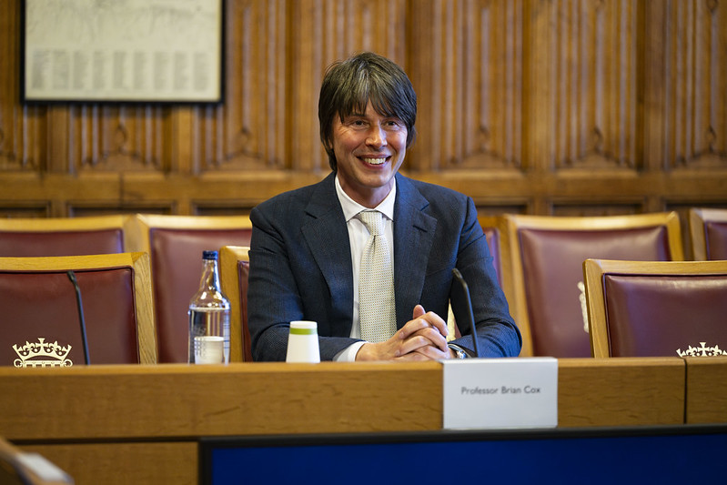 Professor Brian Cox speaking at a House of Lords committee session, seated at a panel desk with a nameplate and microphone, as part of the UK Engagement with Space inquiry.